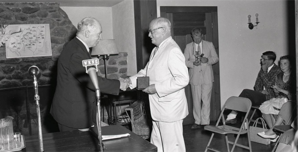Black and white photo of a the first Medal ceremony in 1960. Thornton Wilder, holding the Medal in his left hand, is shaking the hand of James Johnson Sweeney.