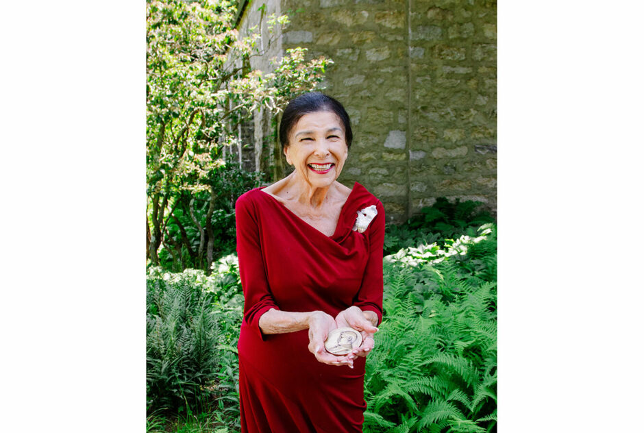 Alanis Obomsawin joyfully holds her medal in her hands. She is wearing a red dress and a silver brooch and standing in front of a stone building surrounded by greenery