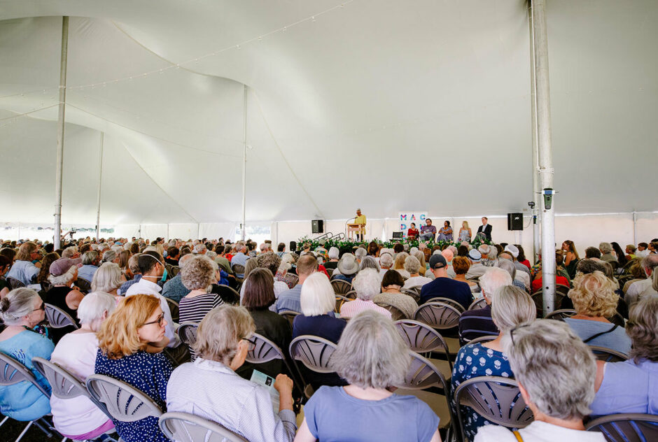 A crowd of over 1,000 gathers under a large white tent to hear speeches and watch Ms. Obomsawin receive her Medal. Sitting in rows of chairs, they all face the stage where a woman stands at a podium