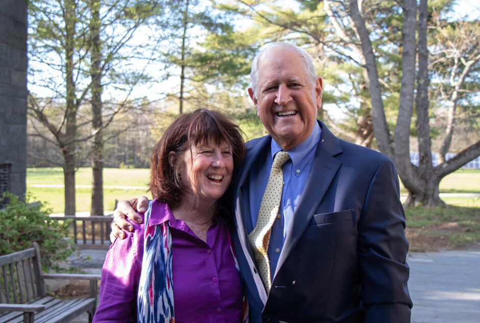 A man and a woman pose together for a photo outside of the library. The man's arm is around the woman and they are both smiling brightly. It is a sunny, spring evening.