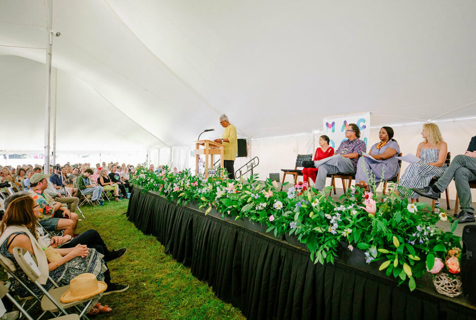 Under a large, white tent, over 1000 people sit in chairs facing a stage. Six people are on the stage awaiting their turn to speak to the audience. A row of flowers lines the edge of the stage and one woman stands at the podium