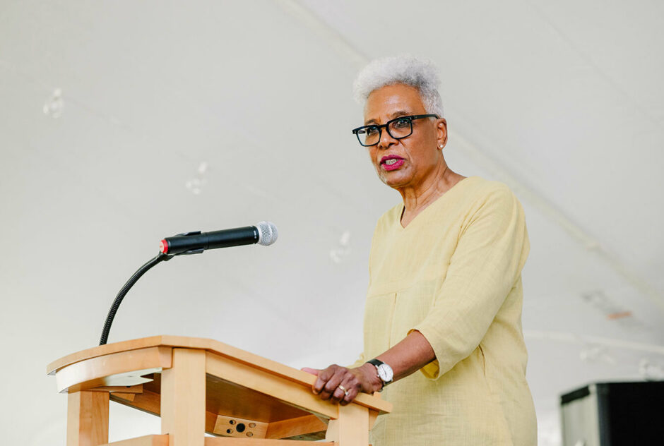 Madam Chairman of the Board Nell Painter stands at a podium and speaks to the crowd. She is centered in the image with only the white vinyl of the tent behind her.