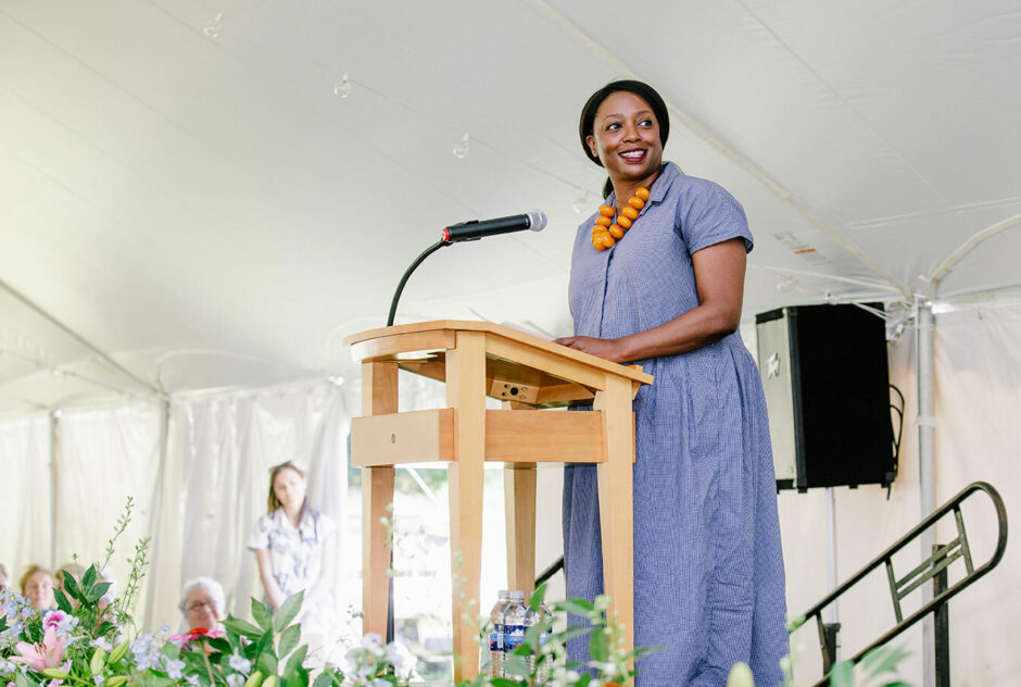 Executive Director Chiwoniso Kaitano stands at a podium and speaks to the crowd. She is centered in the image with only the white vinyl of the tent behind her. Along the bottom of the image, you can see a row of flowers