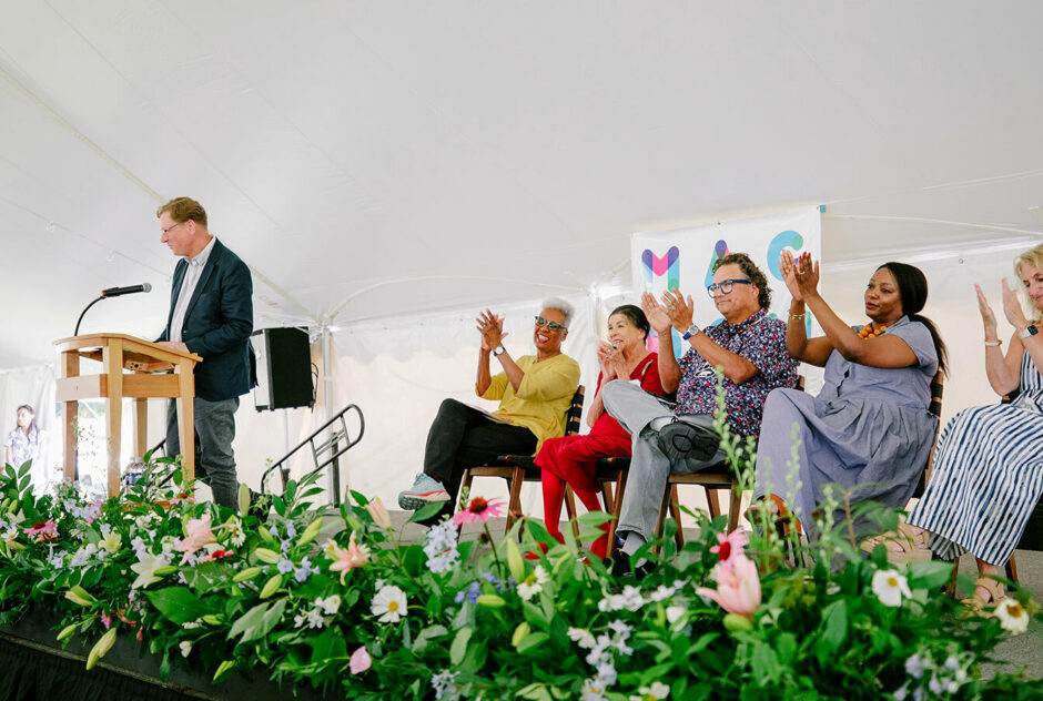 On a small stage under a large white tent, the ceremony speakers sit in a row, one of them, Resident Director David Macy, stands at the podium and speaks to the audience. The front of the stage is lined with flowers.