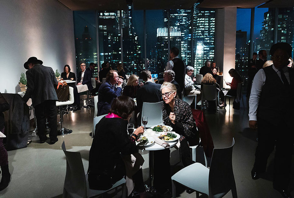 Event attendees mingle, eat and drink in a room with large windows overlooking the city at night