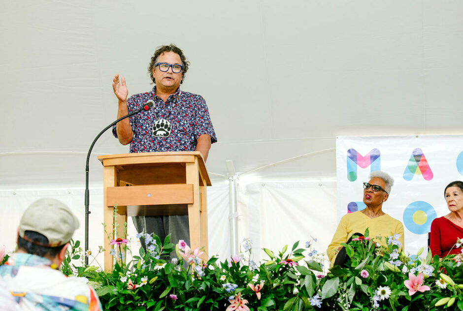 Jesse Wente, a tall man, stands at a podium, gesturing with his hand, and addresses the audience. To his right on stage, two women sit in chairs waiting their turn to speak to the crowd. The front of the stage is lined with a row of flowers.