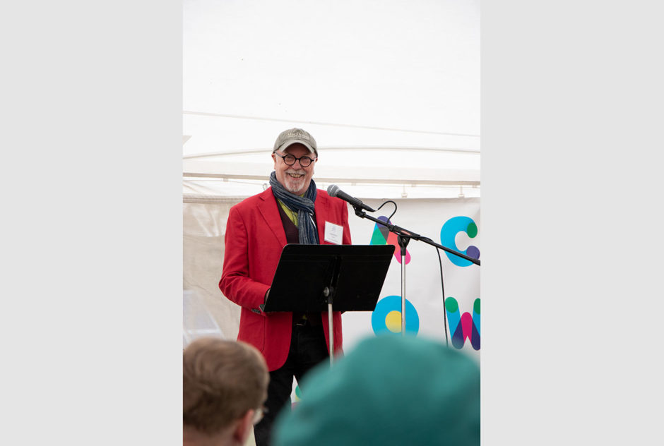 Fellow, board member, and evening emcee Dan Hurlin offers a local perspective of MacDowell and introduces the presenters at the New Hampshire Benefit in May 2022. He stands on stage and wears a red jacket,