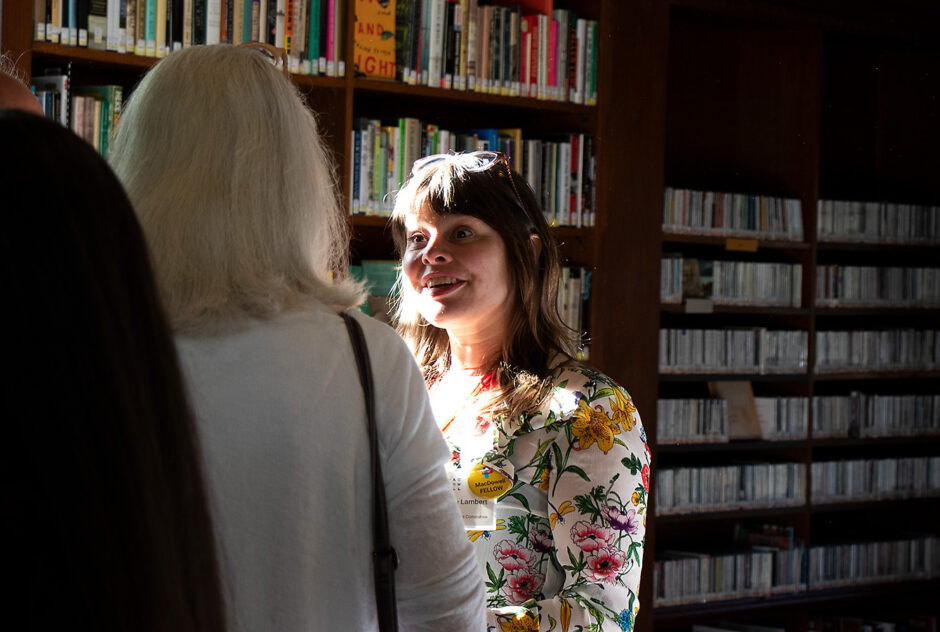 Two women engage in conversation in the library. One woman faces the camera and is shrouded in bright sunlight that is coming through a large window. The light illuminates her smiling face and colorful floral shirt.