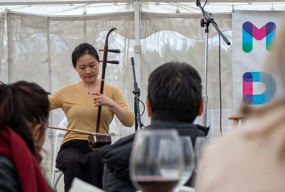 Composer and Fellow Jing Wang plays the erhu (a thin, stringed instrument played with a bow) for the crowd under the tent at the New Hampshire Benefit in May 2022. She looks down at the instrument as she plays.