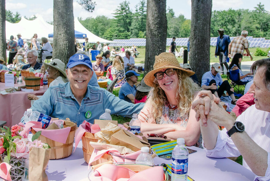On a sunny afternoon people gather at tables and on picnic blankets to eat lunch. Three people, seated at a table with pastel colored linens and flowers, smile for the camera.