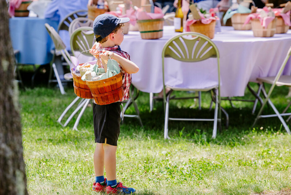 A little boy holding two picnic baskets stands and looks toward the other picnickers. Behind him is a table set with pastel linens.