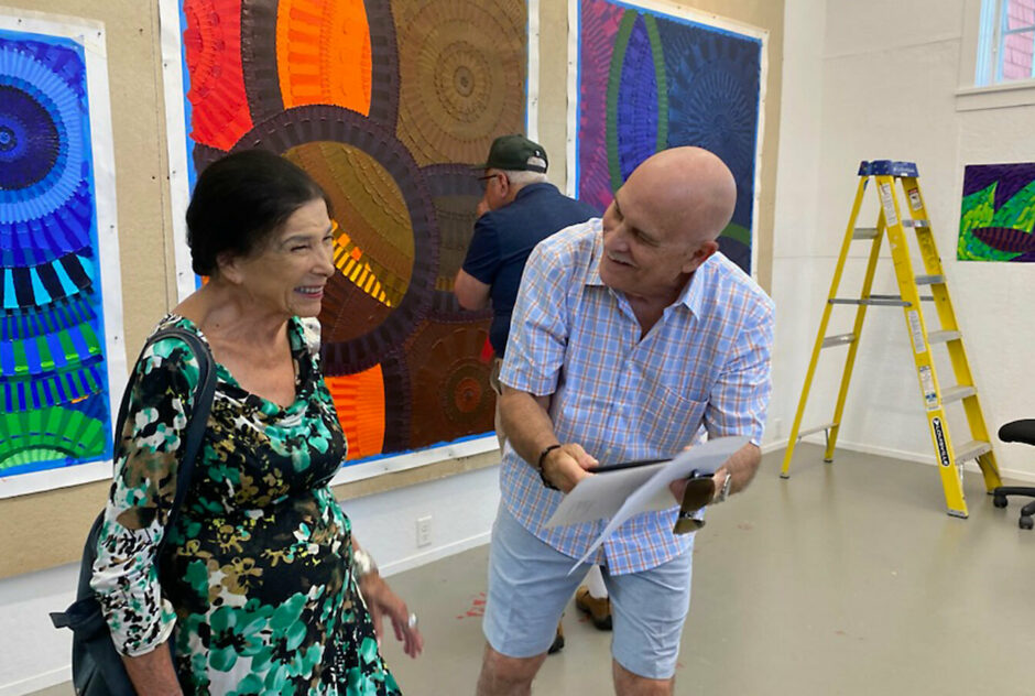 A man and a woman talk and laugh together while inside an artist studio. There are large, boldly colored artworks hanging on the wall behind them.