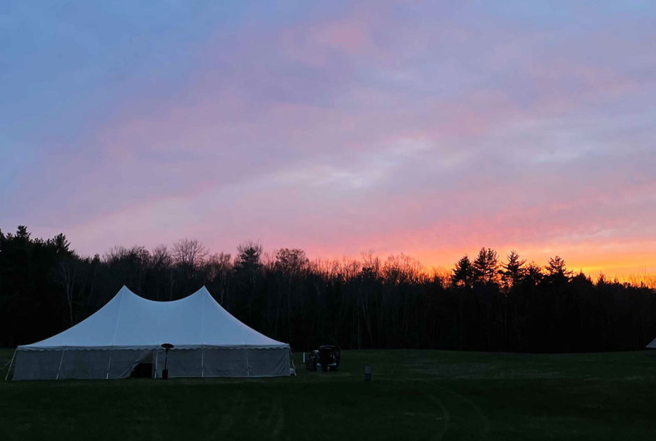 The sun sets over the Medal Day tent, glowing purple and orange at the end of the evening of the New Hampshire Benefit in May 2022. The trees are silhouetted against the sky.