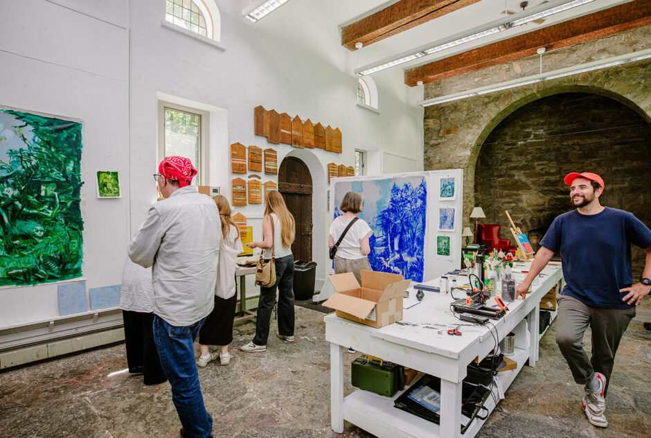 A small group of people tour an artist studio. They all have their backs toward the camera as they study the artwork hung on the walls. The artist stands at a table in the center of the room smiling brightly