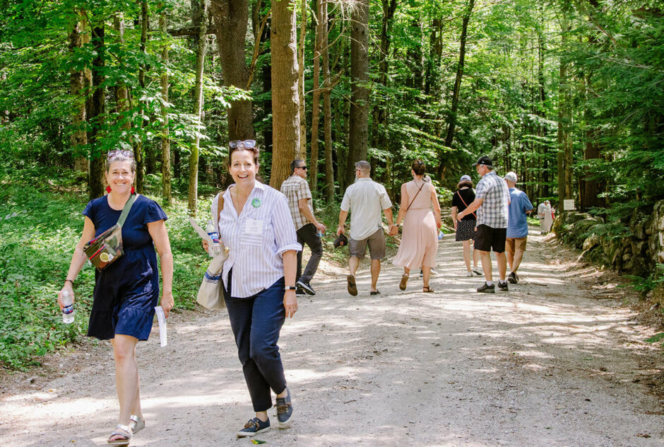 Groups of people walk up and down a dirt road that leads to artist studios. The road is lined with thick, green forests on each side.