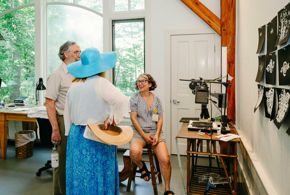 Two people stand next to an artist who sits on a stool. They are talking and laughing with one another as they admire the artists work that is hung on the walls around the studio.