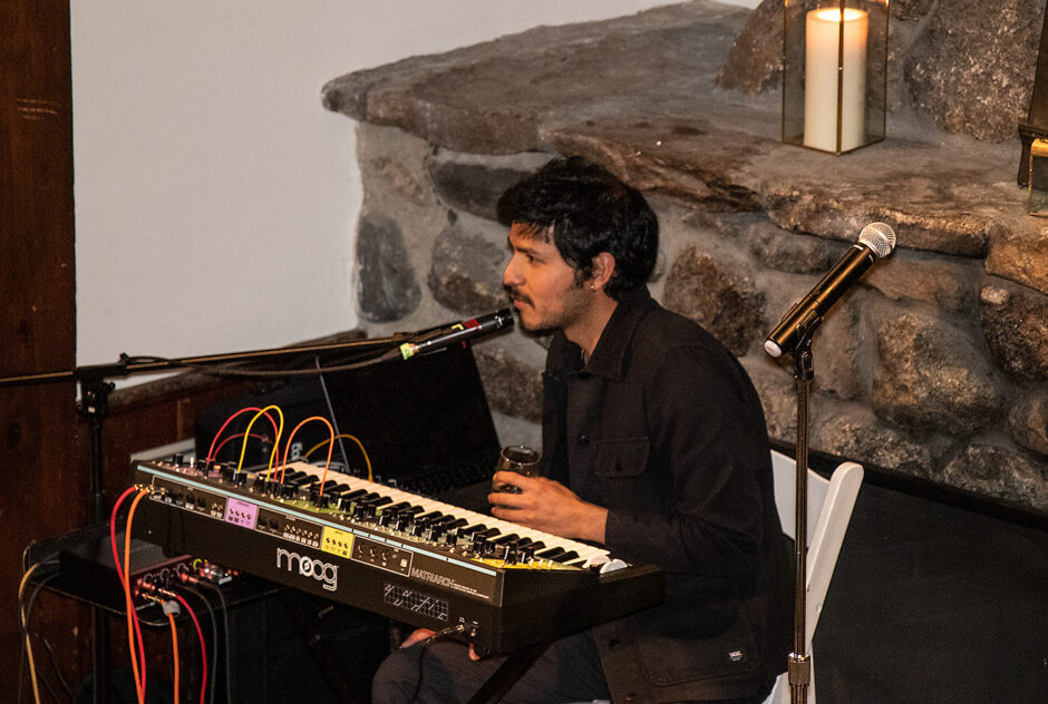 A man sits at an electronic piano, speaking to the audience before beginning his performance. He is on a small stage in front of a large stone fireplace.