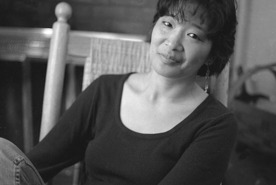 Portrait of Katherine Min in Chapman Studio in 2004. She smile directly into the camera while sitting with one leg up.