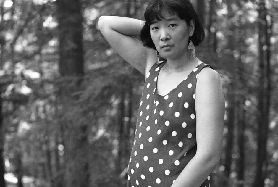 Katherine Min during her residency in 1995. She is wearing a polka dot dress and standing in the woods. One of her arms is held behind her head, the other at her side.
