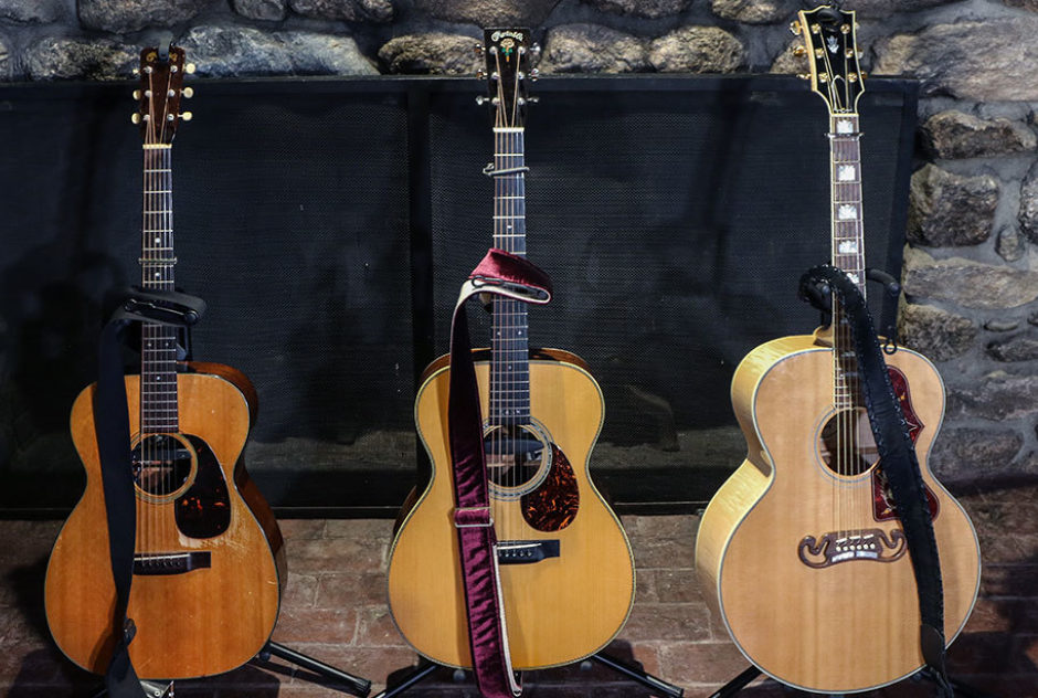 Three acoustic guitars sit on their stands in front of a large fireplace awaiting soundcheck