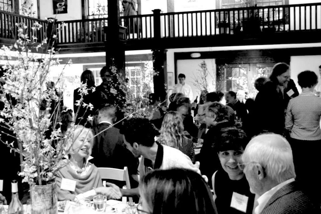 Black and white photograph of MacDowell supporters gathered in the main hall at MacDowell at the New Hampshire Benefit in 2014. Four people are seated at a table in the foreground with a tall floral arrangement on the left.