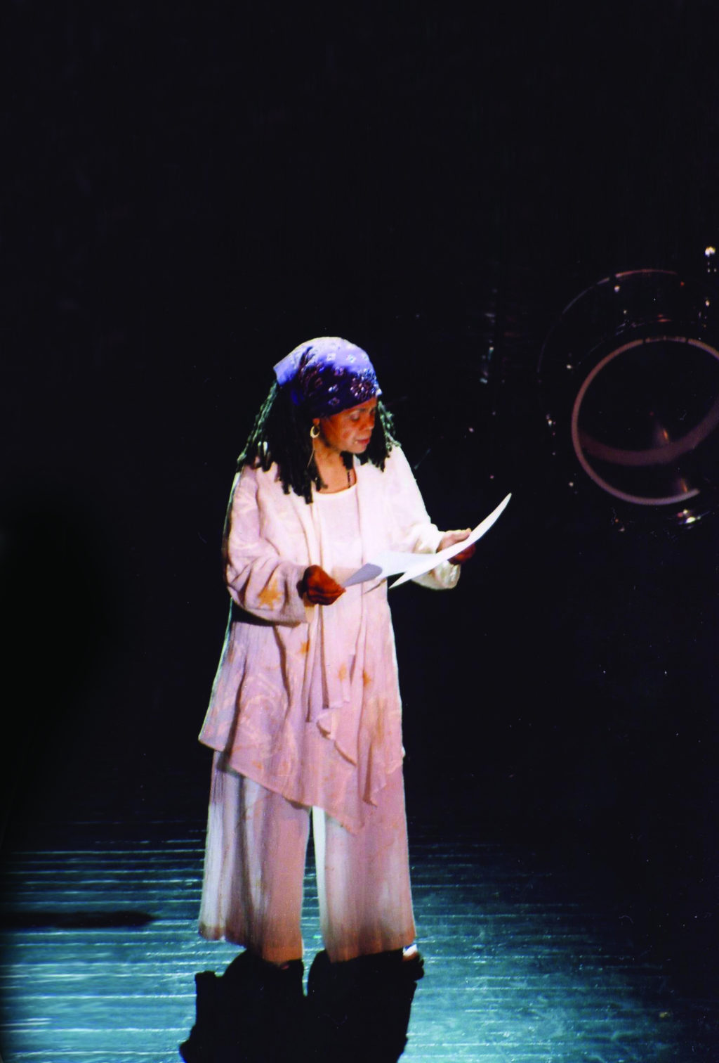Sonia Sanchez, wearing white and a purple hair wrap, is illuminated on stage as she reads off of a paper to the audience