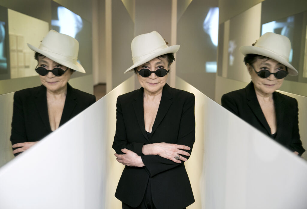 A woman stands in a white fedora, sunglasses, and black suit with her arms crossed, with her reflection mirrored on both sides of her body