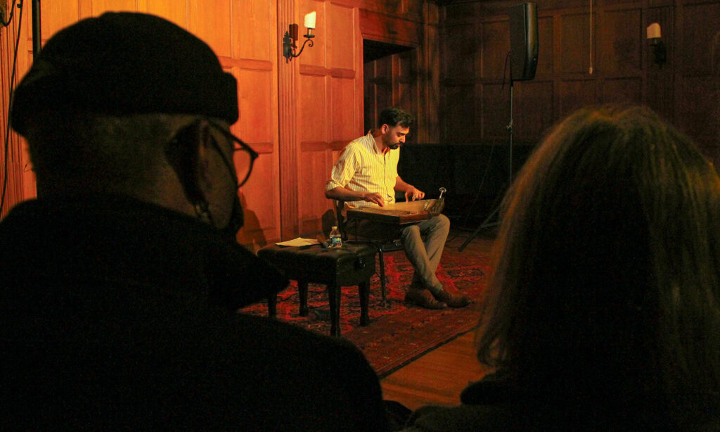 A man sits at the front of a room, facing an audience. He is playing a stringed instrument that rests on his lap. The lighting in the room is warm and inviting.
