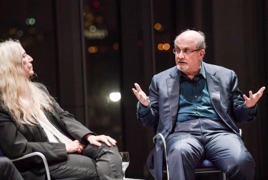Salman Rushdie sits with a woman on a stage. He is speaking to her and is gesturing with his arms