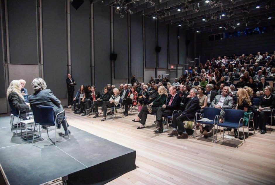Three people sit together on a stage for a panel discussion. In front of them, a large audience who listen eagerly