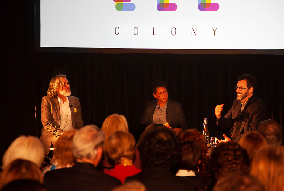 Three people sit in chairs on a stage for a panel discussion, a crowd looks on