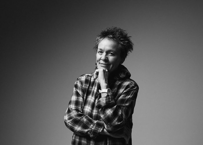 Laurie Anderson poses for a portrait against a plain grey background. Her chin resting in her hand