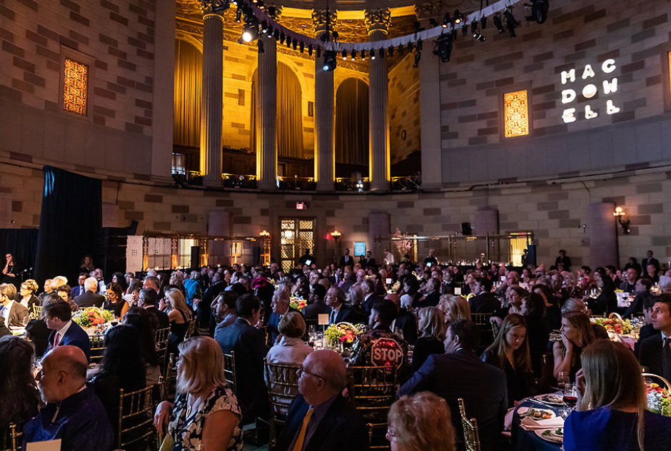 Event attendees, seated at their tables, enjoy performances, speeches, and other pieces of the program. They are in a large, cathedral ceilinged ballroom