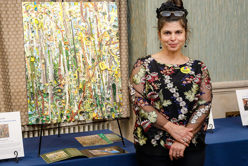 A Fellow stands beside her painting. The colorful, abstract painting is resting on an easel on top of a table