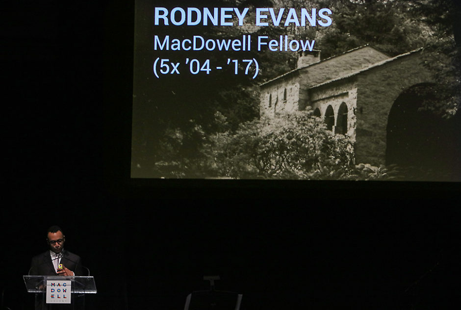 A man speaks into a microphone on a dark stage. Above him is a large screen with the name Rodney Evans and an image of a MacDowell studio on it.