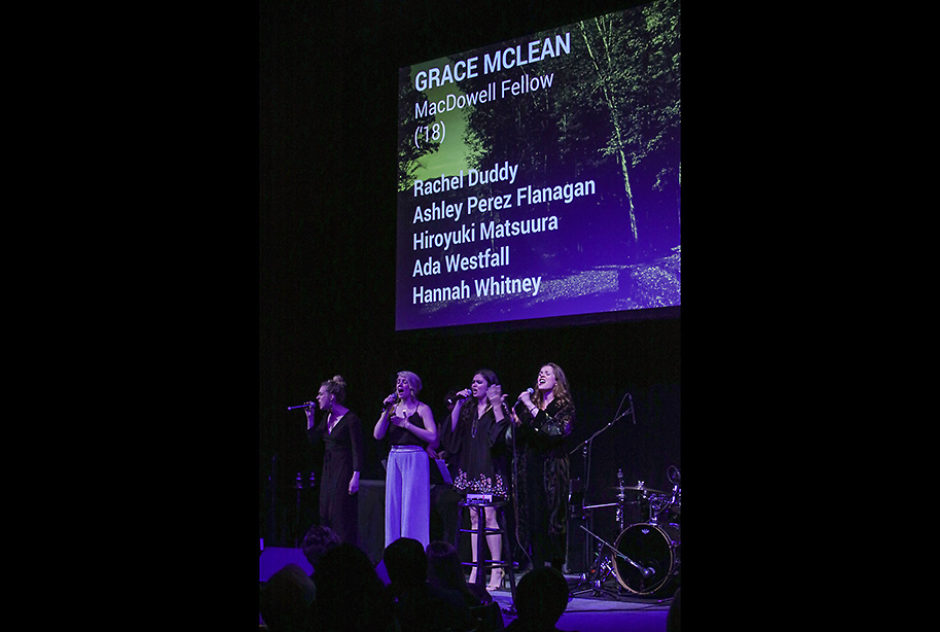 Four performers on a dark stage. Above them is a large screen with the name Grace Mclean and information about the artist.