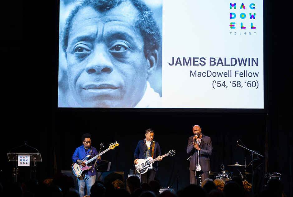 Three performers stand on a dark stage, singing and playing guitars. Above them is a large screen with a portrait of James James Baldwin
