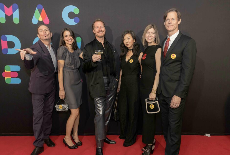 Five people wearing colorful festive clothing stand on a red carpet in front of a black step and repeat that says MacDowell