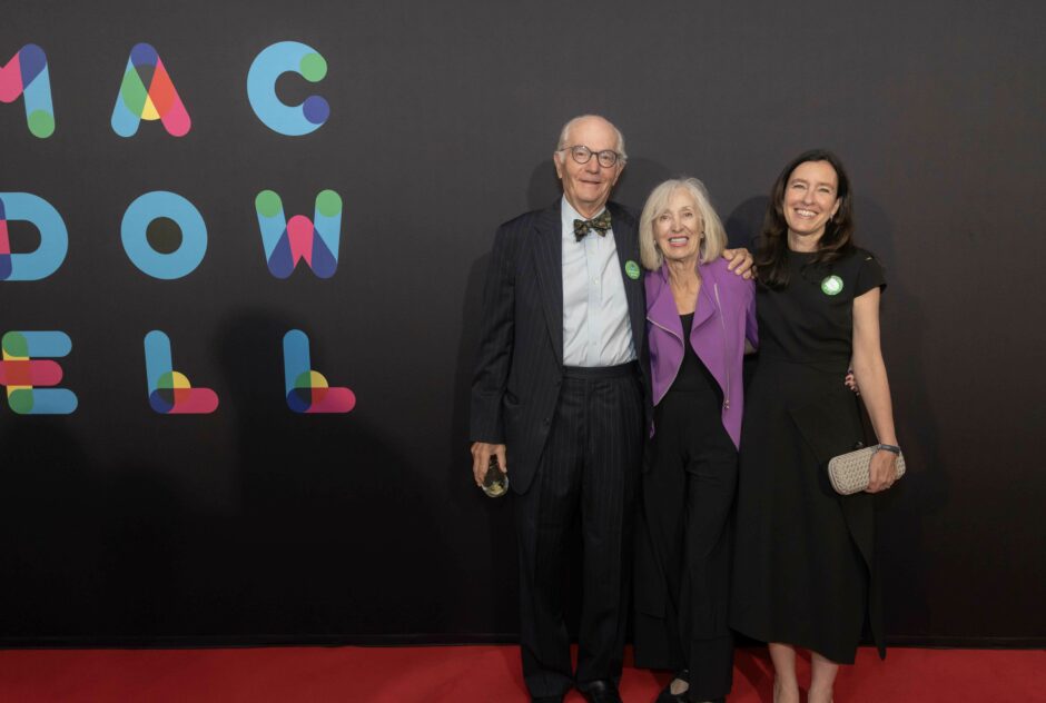 A man in a suit, a woman in a purple jacket, and a woman in a black dress stand on a red carpet against a black step and repeat that says MacDowell