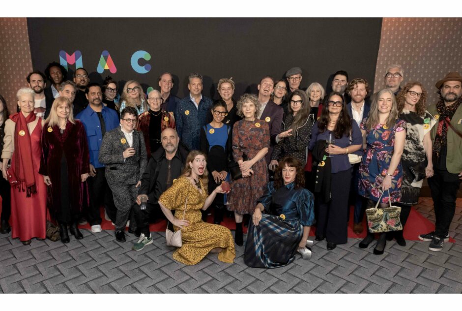 A group of people about 30 people wearing colorful, festive clothing pose in front of a black step and repeat that says MacDowell