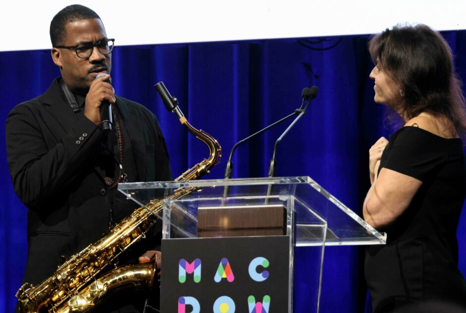 A man carring a saxophone speaking into a microphone on stage while a woman in a black dress faces him