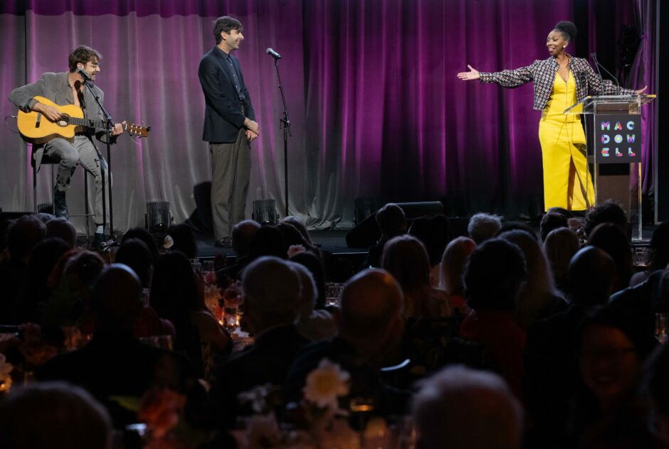 A man holding a guitar sits on stage while a man in a gray suit and a woman in a yellow dress stand on stage. A crowd watches the stage.