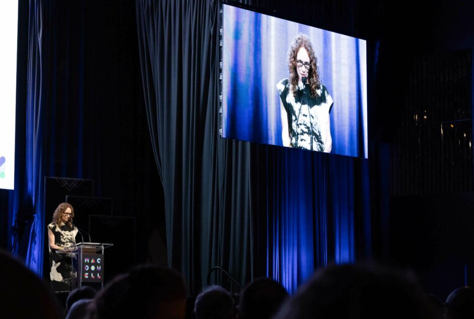 A woman speaks on stage into a microphone while screens behind her project live images of her speaking