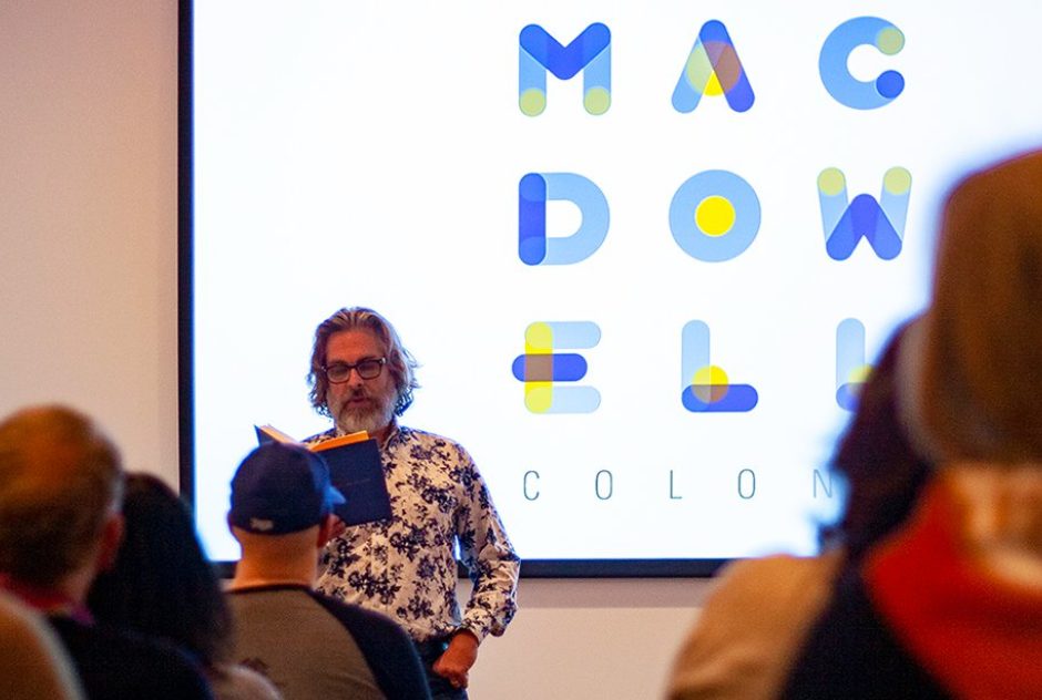 Michael Chabon reads from his book "Bookends" to an audience from the front of the room. He stands in front of a projector screen that features the MacDowell logo