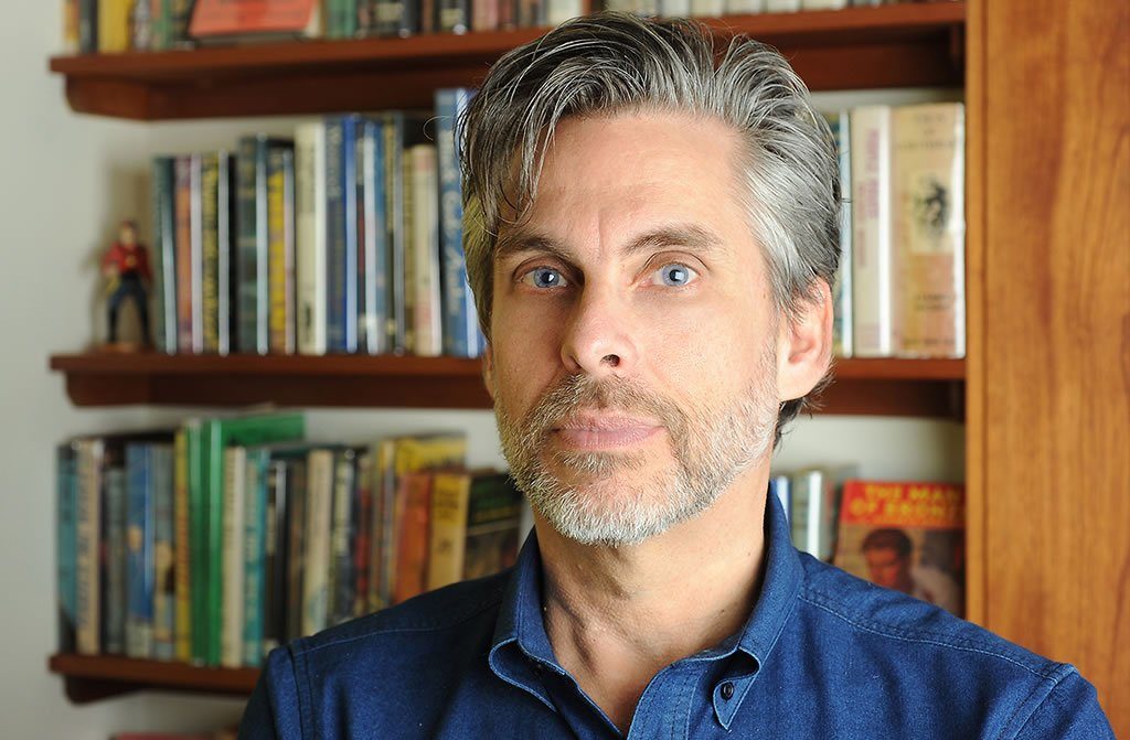 Headshot of Michael Chabon. He is wearing a blue shirt and is posing in front of a book case.