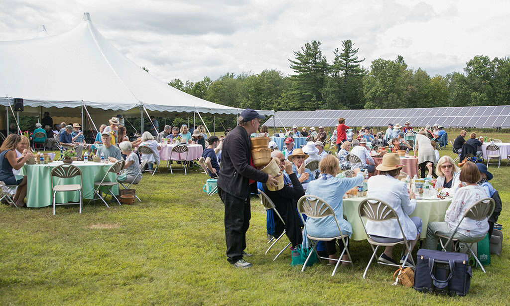 Picnickers sit around tables, enjoying their lunch on Medal Day. A large tent is in the background