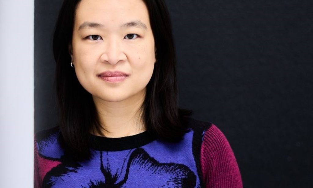 Portrait of composer Tonia Ko. She leans against a wall in a brightly colored floral sweater and smiles softly