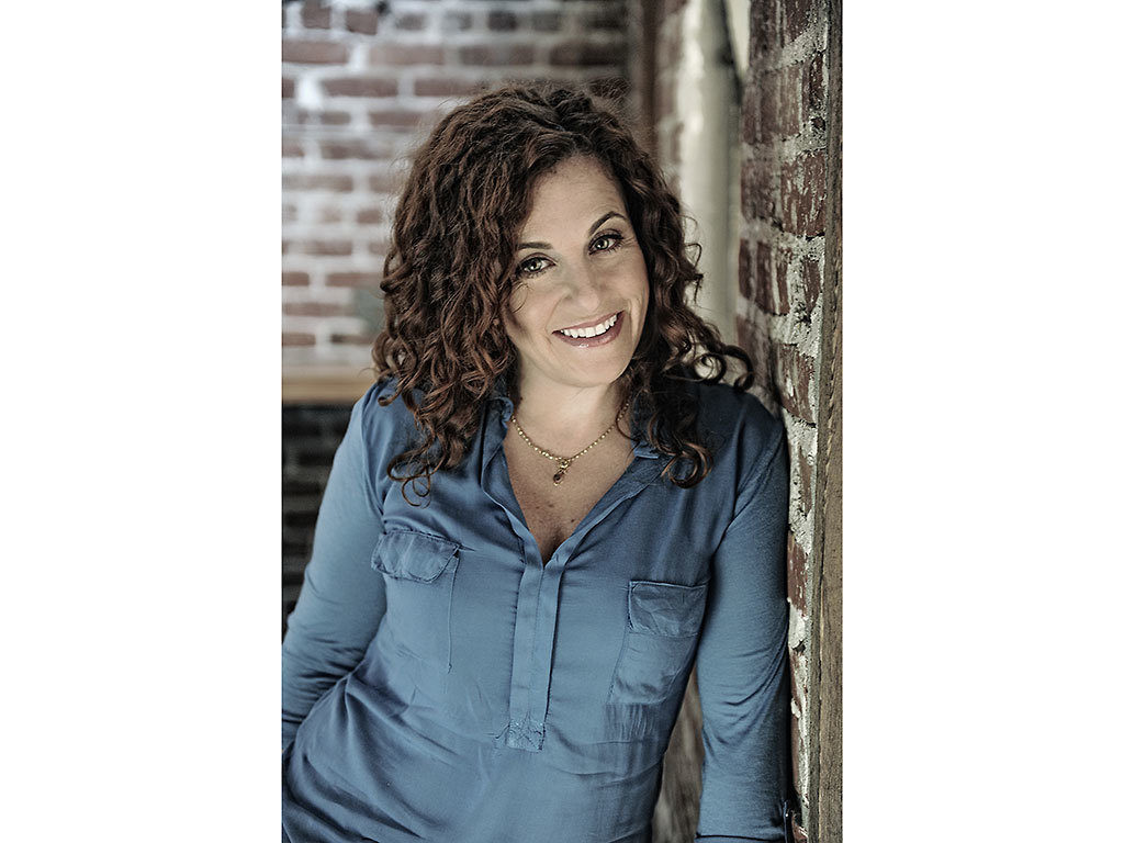 Portrait of Fellow Ayelet Waldman. She is wearing a blue shirt, smiling, and leaning against a brick wall