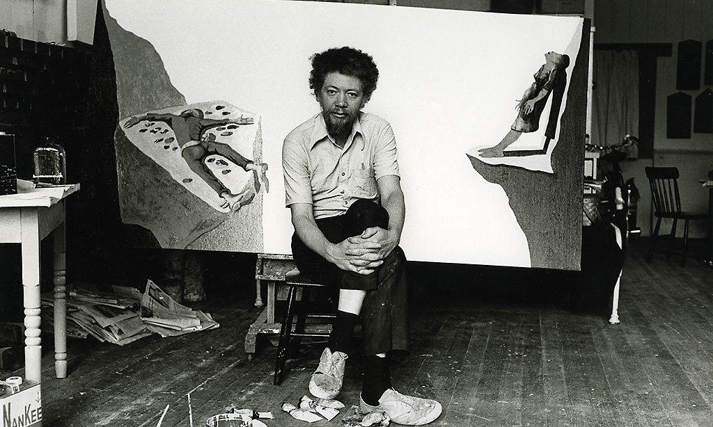 Portrait of Benny Andrews in 1975. He is sitting on a low chair and hugging his knee