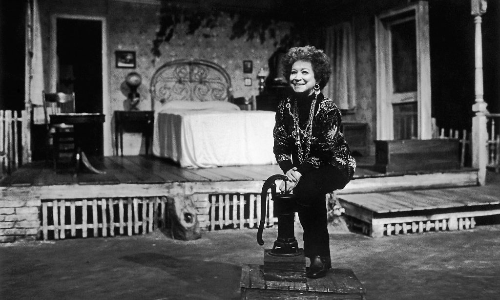 Alice Childress sitting on a stool on the set of one of her plays.